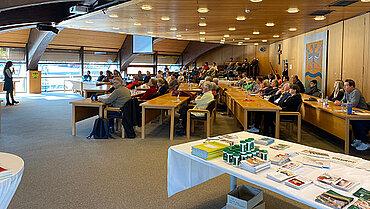 Viele Menschen sitzen an Tischen und schauen nach vorne, wo eine Frau steht und in ein Mikrofon spricht.