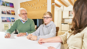 Ein Junge sitzt mit einem erwachsenen Mann und einer erwachsenen Frau an einem Tisch in einer Wohnung.
