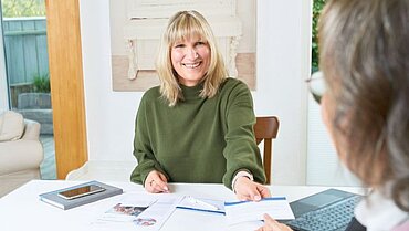 Eine Pflegeberaterin sitzt mit einer Frau am Tisch und gibt ihr eine Unterlage. Die Pflegeberaterin lächelt.