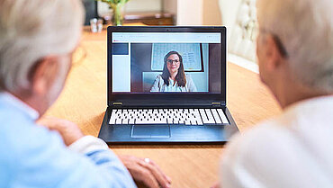 Zwei grauweiß behaarte Personen sitzen mit dem Rücken zur Kamera. Zwischen ihnen ist ein aufgeklappter Laptop zu sehen und auf dem Bildschirm eine lächelnde Frau mit ebenfalls grauen Haaren, die ein Headset trägt, das zugehörige Mikrofon vor ihrem Mund.
