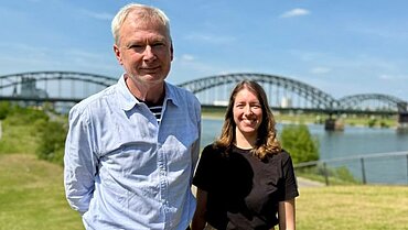 Pflegeberater*innen Olaf Arps und Leonie Hausmann am Rhein in Köln. Es ist ein sonniger Tag. Im Hintergrund ist die Südbrücke zu sehen.