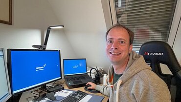 Ein Mann sitzt an einem Schreibtisch vor einem Laptop und einem Bildschirm und lächelt in die Kamera.