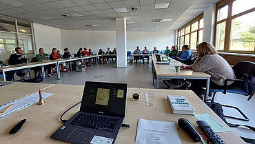 Bild zeigt eine Konferenzsituation, im Vordergrund Laptop und im Hintergrund Menschen