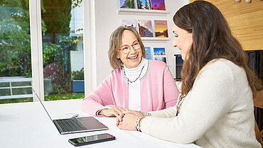 Eine jüngere und eine ältere Frau sitzen an einem Tisch vor einem Laptop, daneben liegt ein Smartphone.