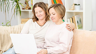 Zwei Frauen sitzen Arm in Arm auf einer Couch und schauen gemeinsam auf ein Laptop.