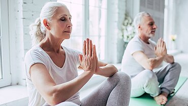 Eine ältere Frau und ein älterer Mann sitzen mit geschlossenen Augen in einer Yoga-Pose auf Matten.