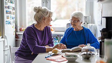 Eine jüngere und eine ältere Frau sitzen gemeinsam an einem Küchentisch und unterhalten sich.