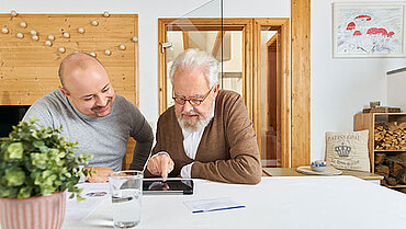 Zwei Männer sitzen an einem Tablet, um sich einen Termin für eine Pflegeberatung zu buchen.