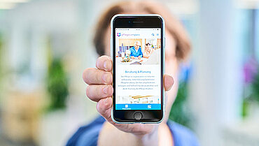 Eine Person hält mit ausgestrecktem Arm ein Handydisplay in die Kamera, auf dem die pflegecompass-App geöffnet ist.