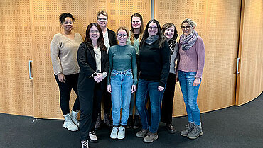 Acht Frauen in legerer Kleidung stehen nebeneinander und lächeln in die Kamera. Sie befinden sich in einem Raum, der Hintergrund ist eine Schrankwand.