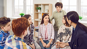 Sechs Kinder und eine erwachsene Person sitzen im Kreis und unterhalten sich.