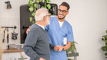 Ein Pfleger stützt mit einer Hand einen älteren Mann, der in der anderen Hand einen Gehstock hält.