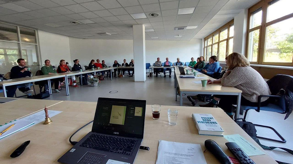 Bild zeigt eine Konferenzsituation, im Vordergrund Laptop und im Hintergrund Menschen